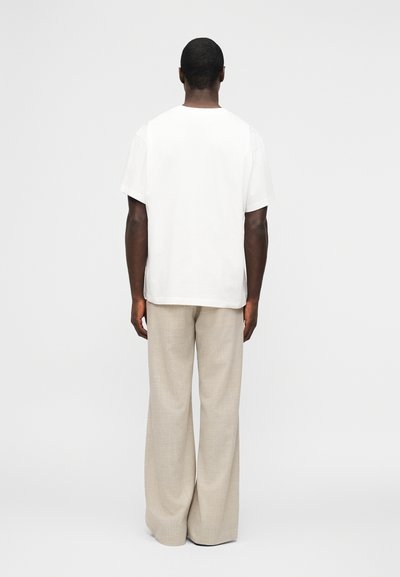 Άνδρας που στέκεται πλάτη με λευκό κοντομάνικο πουκάμισο και χαλαρό μπεζ παντελόνι μπροστά σε απλό ανοιχτό φόντο.