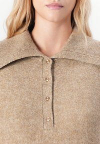 Beige ullblandingsgenser med stor krage, med en knappestolpe og fem lyse knapper. Myk, teksturert stoff.
