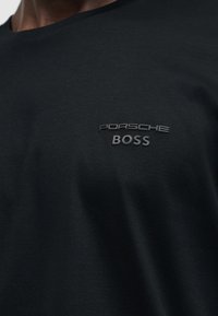 Μαύρο πουκάμισο με ανάγλυφο λογότυπο "Porsche BOSS" στο πάνω μέρος του στήθους, φορεμένο από άτομο με σκούρο χρώμα δέρματος.