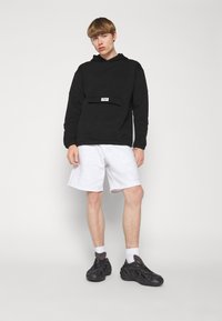 Felpa nera con cappuccio e tasca frontale, decorata con un logo bianco, abbinata a pantaloni corti grigi chiari e scarpe da ginnastica nere. Il modello è in piedi su uno sfondo neutro.