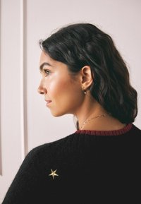 Suéter negro con cuello acanalado rojo, que presenta un bordado de una estrella dorada en la parte de atrás. El modelo tiene el cabello oscuro y ondulado y lleva pendientes de aro.