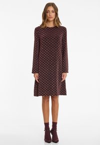 Knee-length dress in dark burgundy with a diamond pattern. Long sleeves, round neckline, and soft texture. Paired with matching socks and heels.