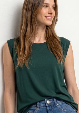 Frau lächelt, trägt ein dunkelgrünes ärmelloses Oberteil und blaue Jeans, steht vor einer hellen Wand und schaut nach rechts.