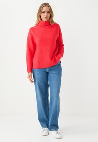 Koralowy sweter z golfem wykonany z miękkiego materiału, z prążkowanymi mankietami i dołem, w połączeniu z niebieskimi dżinsami o luźnym kroju i białymi trampkami.