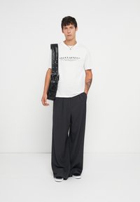 Λευκό μπλουζάκι με μαύρο λογότυπο, σκούρο παντελόνι με φαρδιά πόδια και μαύρη τσάντα χιαστί. Αξεσουάρ περιλαμβάνουν ασημένιο κολιέ αλυσίδα.