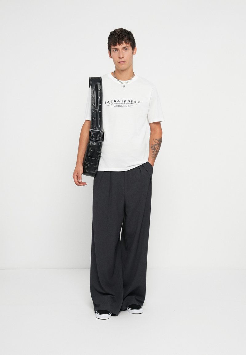 Λευκό μπλουζάκι με μαύρο λογότυπο, σκούρο παντελόνι με φαρδιά πόδια και μαύρη τσάντα χιαστί. Αξεσουάρ περιλαμβάνουν ασημένιο κολιέ αλυσίδα.