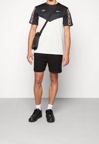Man wearing black and white Nike sports t-shirt, black shorts, black sneakers, white socks, and a black crossbody bag against plain background.