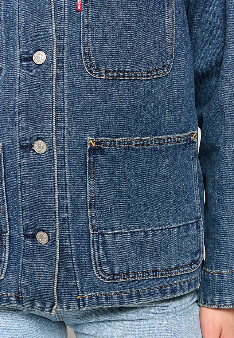 Chaqueta de denim de color azul medio, con dos grandes bolsillos frontales, botones de metal y costuras blancas contrastantes.
