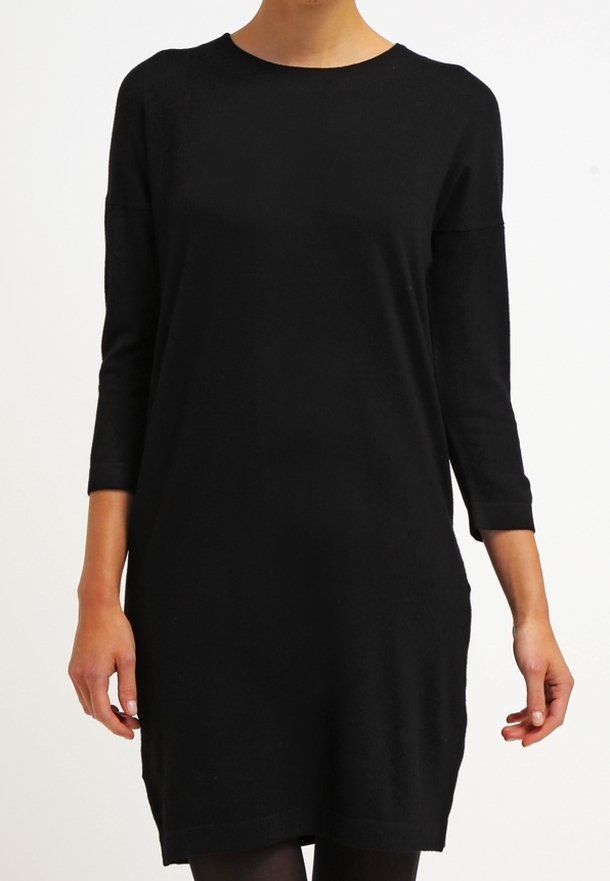 Femme portant une robe noire simple arrivant au genou avec des manches trois-quarts, debout devant un fond blanc uni.