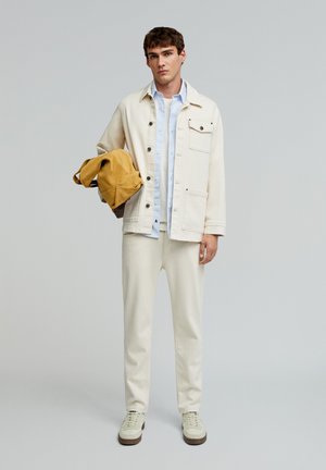 Light beige denim jacket with buttons, paired with a blue striped shirt, beige pants, and off-white sneakers. A brown bag is held in one hand.