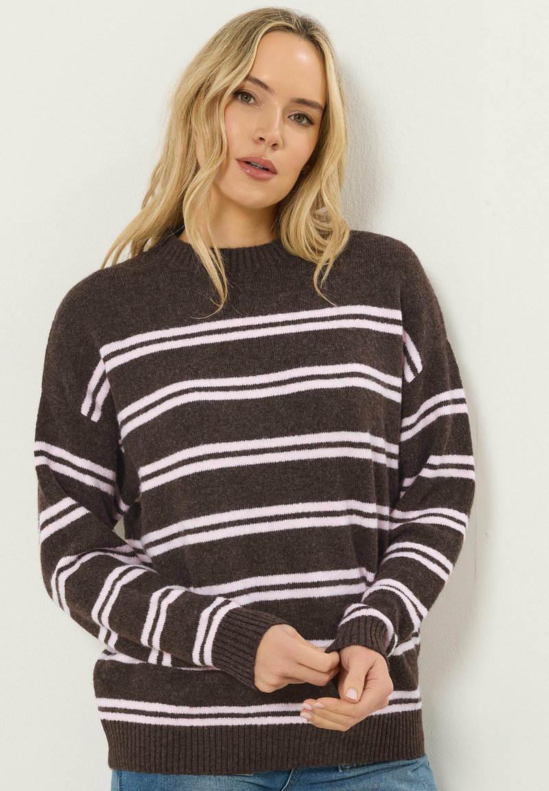 Donna con capelli biondi lunghi che indossa un maglione marrone scuro con strisce orizzontali rosa chiaro e jeans blu, in piedi contro un muro bianco.