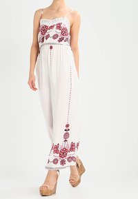 Vitt jumpsuit med röd blomsterbroderi, spetsdetaljer och justerbara spagettiband. Har vida ben och dragsko i midjan.