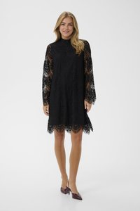 Vrouw staand in een zwarte kanten jurk met lange mouwen en bordeauxrode spitse slingback pumps met hak, glimlachend tegen een effen achtergrond.