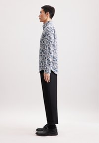 Uomo in profilo che indossa una camicia lunga a maniche blu con motivi floreali, pantaloni neri, calze nere e scarpe eleganti nere su uno sfondo semplice.