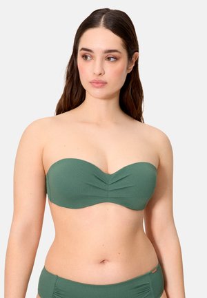 Joven mujer con cabello largo y castaño, llevando un top de bikini verde sin tirantes y un pantalón alto a juego, contra un fondo claro.