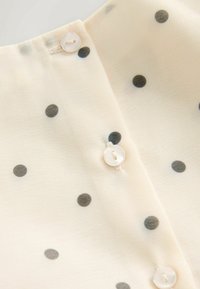 Kremowa bluzka z tkaniny w czarne kropki, z zapięciem na guziki przy szyi i gładką, miękką teksturą.