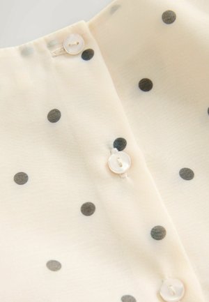 Kremasta bluzica od tkanine s crnim točkicama, koja ima gumbe na vratu i glatku, mekanu teksturu.