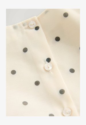 Krämfärgad blus i tyg med svarta prickar, med knäppning vid halsen och en mjuk och slät textur.
