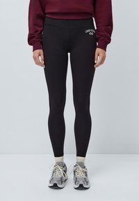  svarte leggings med høy midje og en jevn tekstur; har hvit logo detalj på venstre hofte. Kombinert med grå sneakers og lyse sokker.