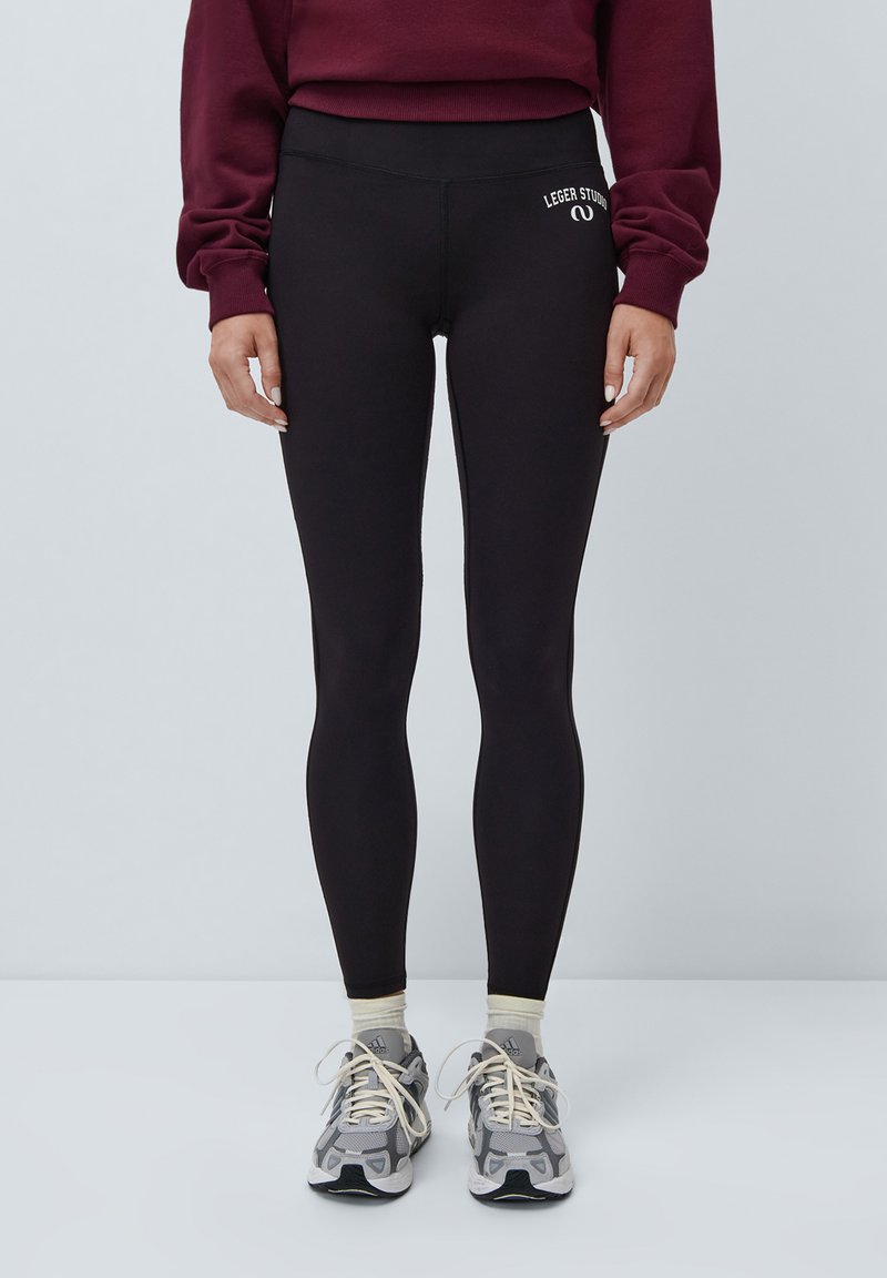  svarte leggings med høy midje og en jevn tekstur; har hvit logo detalj på venstre hofte. Kombinert med grå sneakers og lyse sokker.