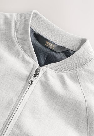 Jachetă bomber gri deschis, cu guler tricotat, fermoar frontal și căptușeală interioară netedă cu modele geometrice. Etichetă cu brand vizibilă.