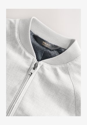 Világosszürke bomberdzseki bordázott gallérral, elülső cipzárral és sima belső béléssel, amely geometriai mintákat tartalmaz. Látható márkajelzés.