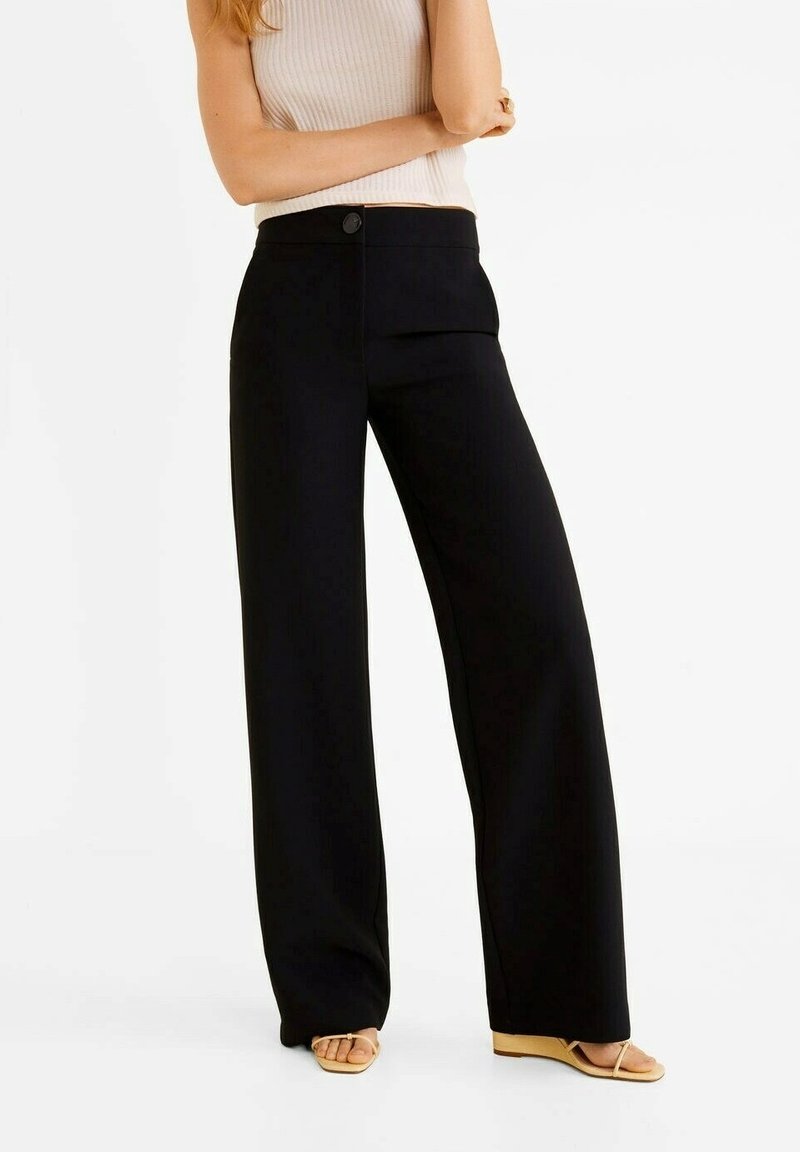 Femme portant un pantalon large noir et des sandales beiges, debout les bras croisés, cadrée au-dessus des épaules.