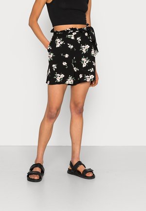 Kvinde iført sorte blomstrede shorts med bindebånd i taljen og sidelommer, sort kort top og sorte sandaler med brede stropper.