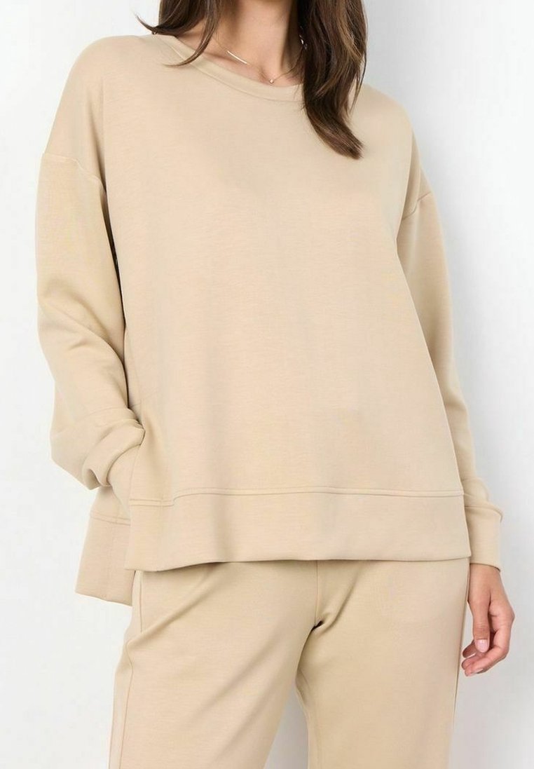Femme portant un sweat-shirt beige oversize et un pantalon assorti, debout avec une main dans la poche sur fond blanc.