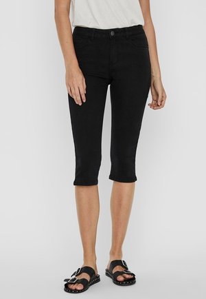 Femme portant un pantalon slim noir longueur genou, un haut blanc et des sandales noires à double boucle et semelles cloutées, se tenant devant un fond uni.