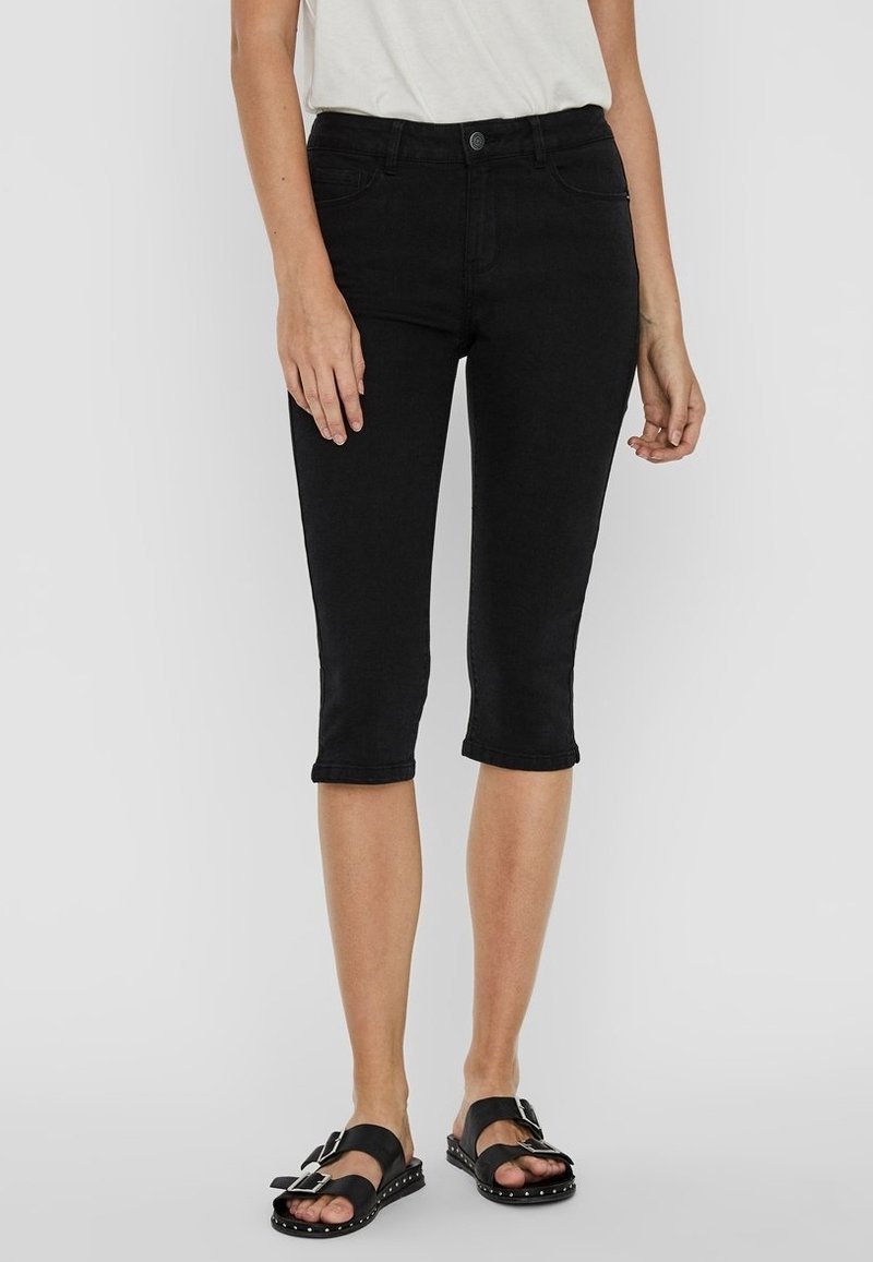Femme portant un pantalon slim noir longueur genou, un haut blanc et des sandales noires à double boucle et semelles cloutées, se tenant devant un fond uni.