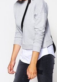 Persona vestida con un sudadera gris claro sobre una camisa blanca y jeans gris oscuro, llevando un bolso de strap negro sobre un hombro.