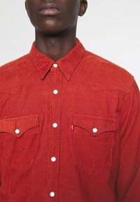 Rostkleurig corduroy overhemd met een knoopsluiting, voorzien van twee borstzakken en witte drukknopen. Geleidende stof met een gestructureerde kraag.