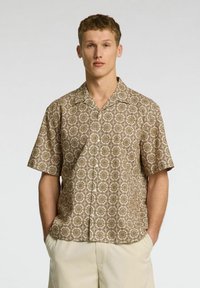 Camicia a maniche corte con colletto in un tessuto beige, caratterizzata da un motivo geometrico ripetuto in marrone e oro. Presenta bottoni sul davanti e una vestibilità comoda.