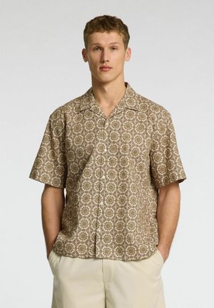 Camicia a maniche corte con colletto in un tessuto beige, caratterizzata da un motivo geometrico ripetuto in marrone e oro. Presenta bottoni sul davanti e una vestibilità comoda.
