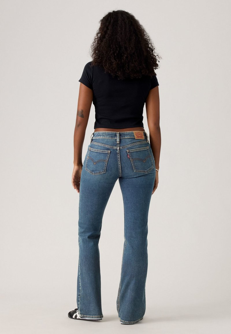 Person with curly hair wearing a black cropped shirt, blue high-waisted flared jeans, and black and white sneakers standing with back facing forward.