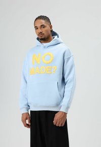 Sweat à capuche bleu clair avec un grand texte jaune "NO MADE ?" sur le devant. Dispose d'une poche kangourou et de poignets côtelés. Associé à un pantalon noir.