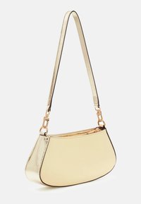 Mała metaliczna złota torebka na ramię z zaokrąglonym dnem, zamkiem błyskawicznym i regulowanym paskiem na białym tle.