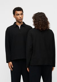 Μαύρο πουλόβερ φούτερ με φερμουάρ στον γιακά, μακριά μανίκια και υφασμάτινη υφή. Διαθέτει άνετη γραμμή και μινιμαλιστικό σχέδιο.