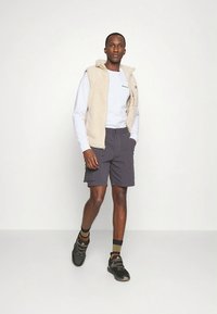 Bézs polár mellény fehér hosszú ujjú ing felett, sötét rövidnadrággal párosítva. Fekete cipők mintás zoknival. Egyszínű háttér.