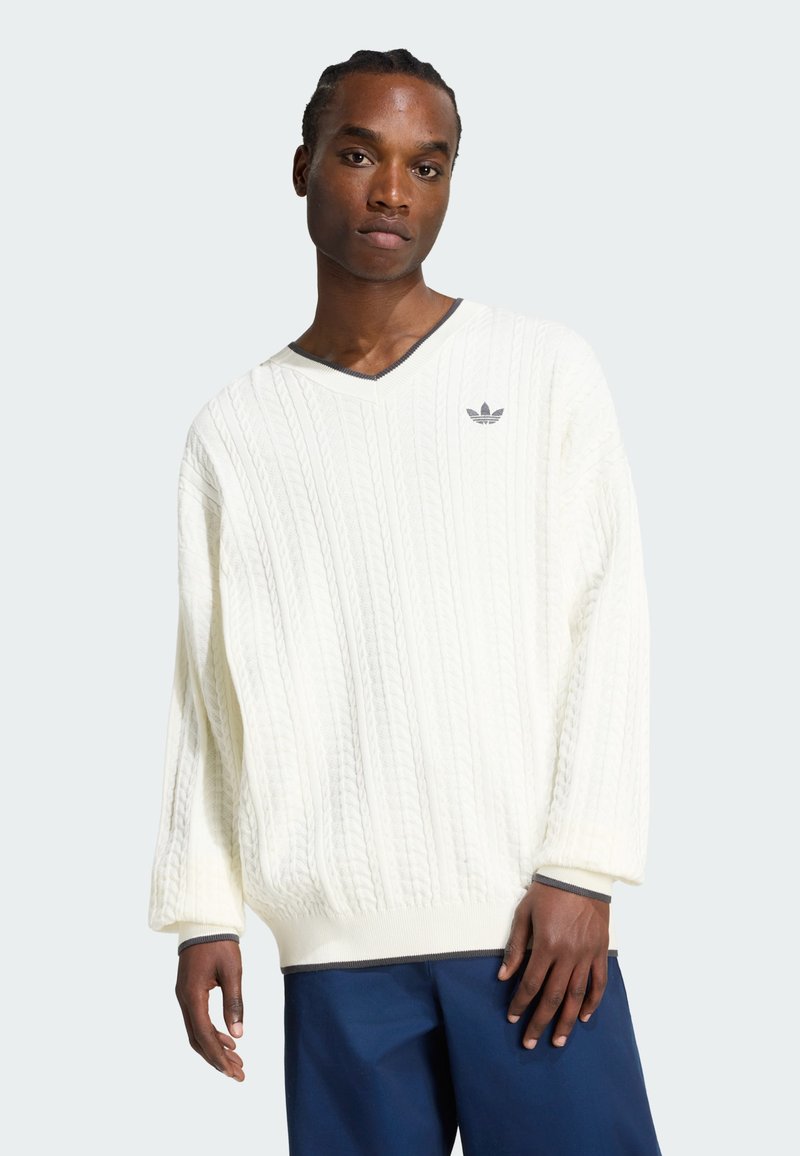 Homme portant un pull blanc à col en V en maille torsadée avec le logo Adidas et un pantalon bleu foncé, debout devant un fond clair uni.
