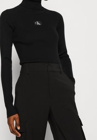 Zwarte geribbelde coltrui met een klein logo-opdruk. Draagt met zwarte broek met zijzakken en een gladde textuur.