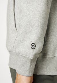 Gray sweatshirt with soft texture, featuring a black embroidered smiley face on the left sleeve and a ribbed cuff.