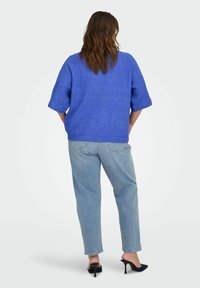 Donna con capelli castani lunghi che indossa un maglione blu a maniche corte, jeans azzurri chiari e scarpe nere con tacchi alti, vista di spalle.