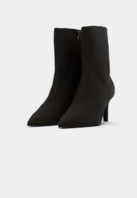 Botas de tornozelo pretas de salto alto com bico fino, feitas de camurça texturizada, com fecho lateral e um corte ajustado.