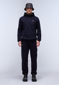 Navy fleece hoodie with half-zip and logo, paired with matching joggers featuring a side pocket. Black bucket hat and shoes complete the look.