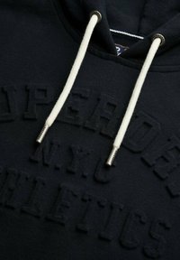 Sudadera navy con lettering en relieve "SUPERDRY N.Y.C ATHLETICS". Cuenta con cordones blancos y herrajes plateados. Textura de tejido suave.