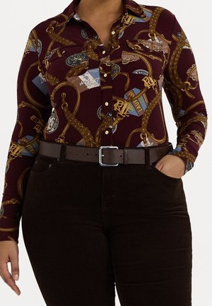 Chemise bordeaux boutonnée avec imprimé corde et emblème, associée à un pantalon marron foncé et une ceinture noire, main reposant dans la poche.