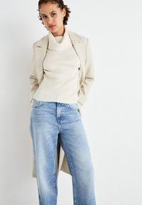 Beige ribota turtlenecka džemperis, apvienots ar gaiši ziliem taisniem biksēm. Modelis valkā krēmkrāsas bleizeri, izceļot neitrālu krāsu paleti.