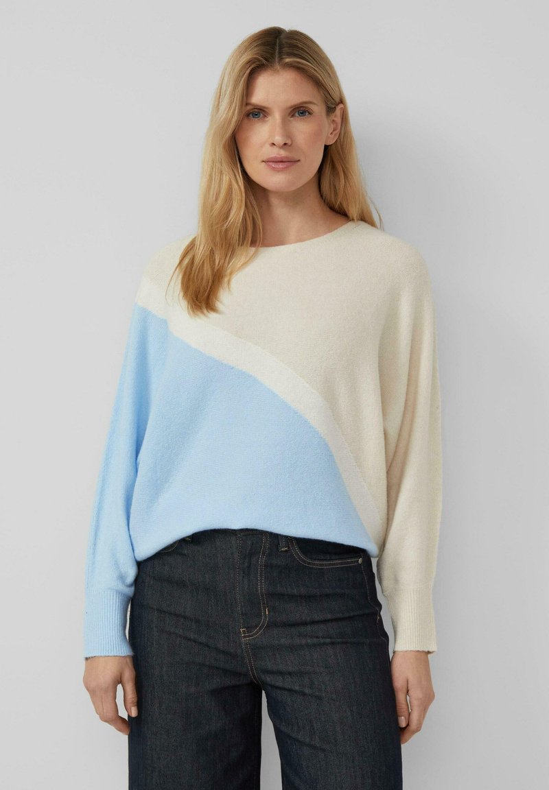 Pull bleu clair et crème avec un design diagonal, une texture douce et des manches longues, associé à un jean en denim foncé.